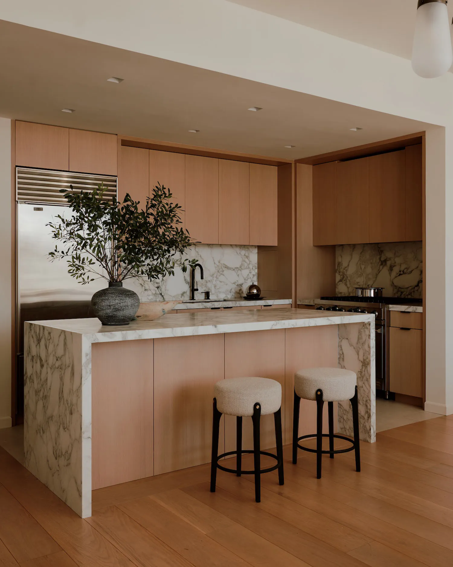 Modern kitchen interior with light oak cabinetry, white and grey marble countertops and backsplash, a large kitchen island, two round fabric bar stools with black legs, a stainless steel refrigerator, and a potted olive tree on the island.