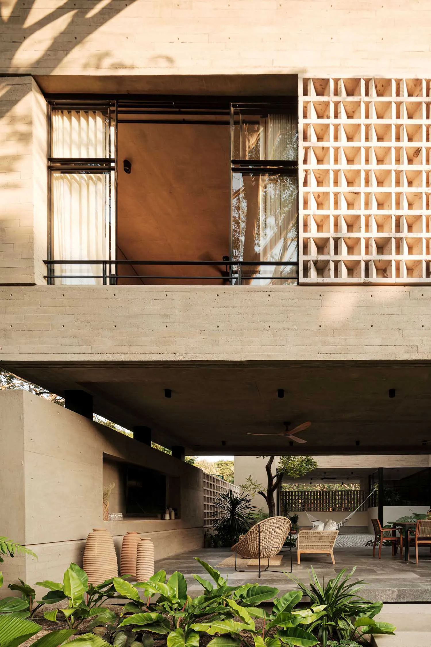 Modern tropical house outdoor courtyard featuring exposed concrete architecture, wooden lattice screen, wicker lounge chairs, and lush tropical plants under warm sunlight.