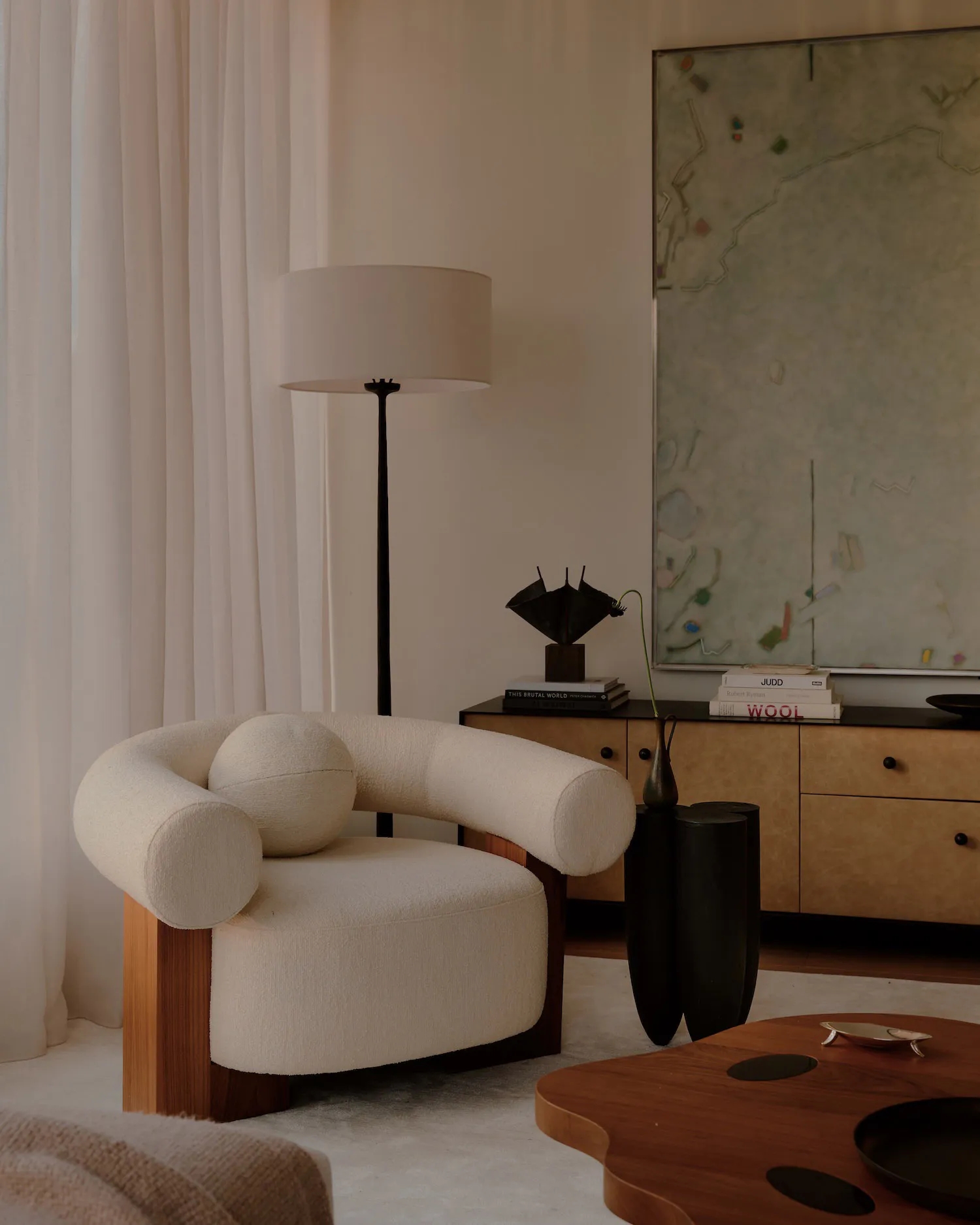 A modern living room featuring a cream-colored curved boucle armchair with a walnut wood base, positioned next to a black floor lamp, dark sculptural side tables, and a wooden console displaying art books and a black metal sculpture, with a large abstract painting on the wall.