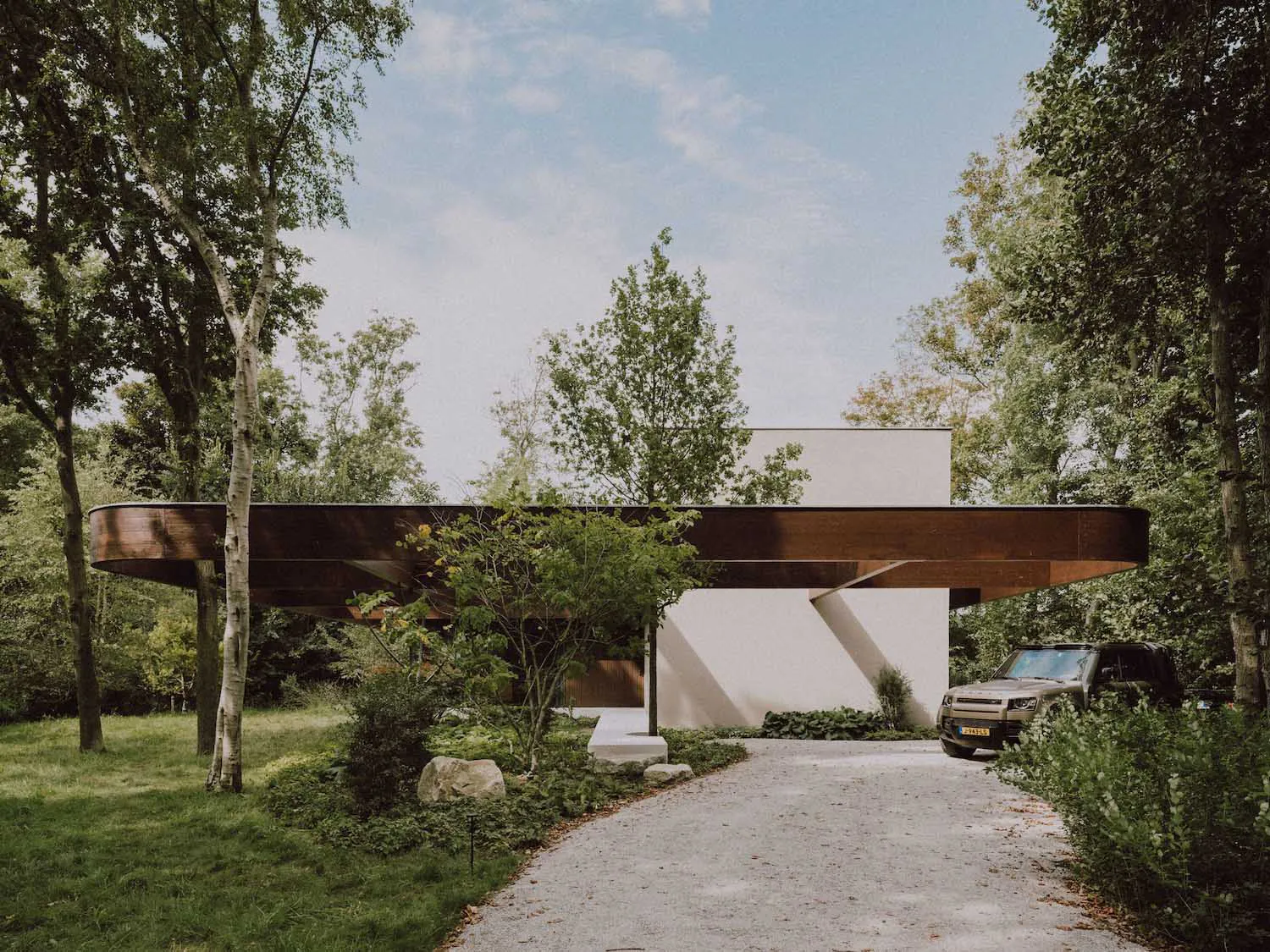 Modern house with a large cantilevered corten steel beam extending over the entrance, surrounded by lush green trees and a gravel driveway with a parked SUV.