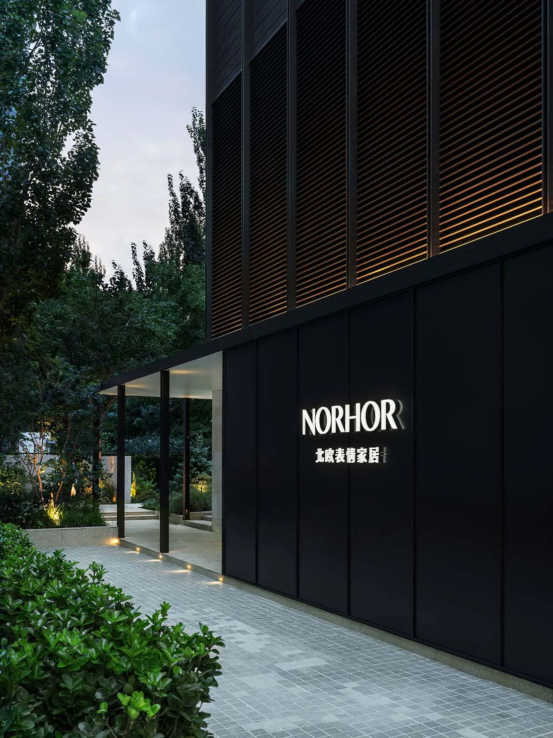 現代黑色建築立面,白色 NORHOR 發光標誌與中文文字,搭配水平木格柵溫暖背光,旁邊是景觀花園步道、樹木與地面燈光,時值黃昏。