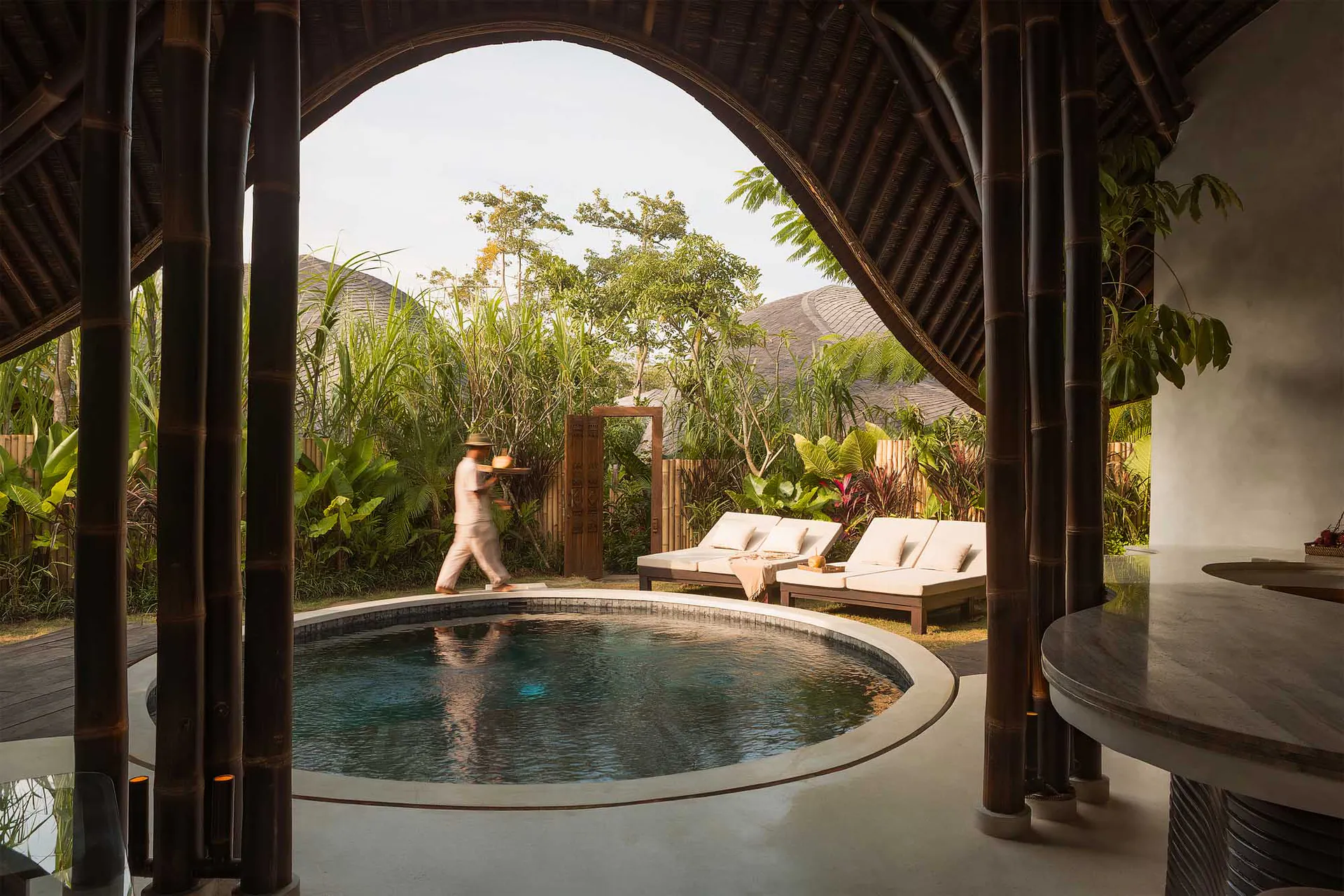 View from inside a dark bamboo pavilion looking out through a large arched opening to a circular swimming pool in a lush tropical garden courtyard, with a staff member carrying a tray and two lounge chairs visible in the background.