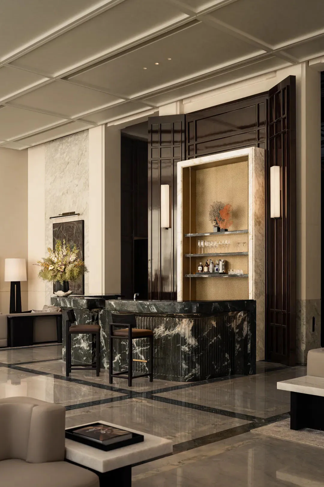 Elegant hotel lobby interior with a curved green marble reception desk, two bar stools, dark wood paneling, and a built-in back bar with glassware and decorative coral.