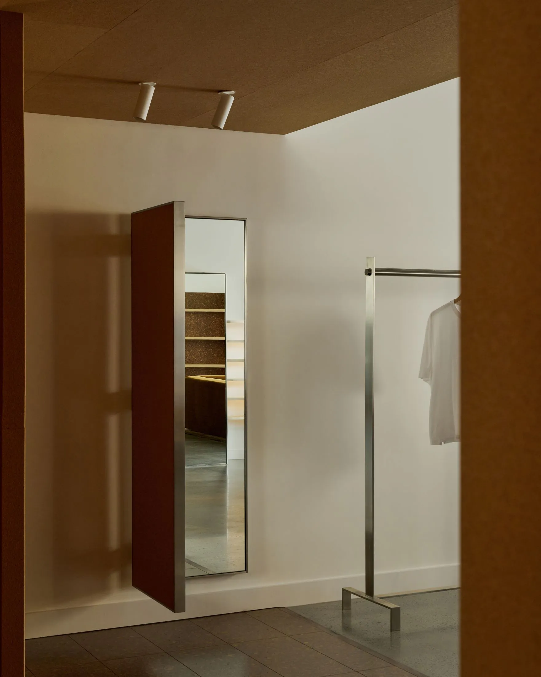 Modern minimalist entryway featuring a large red pivot door with stainless steel frame, concrete terrazzo floor, white walls, and a stainless steel clothing rack with a white t-shirt.