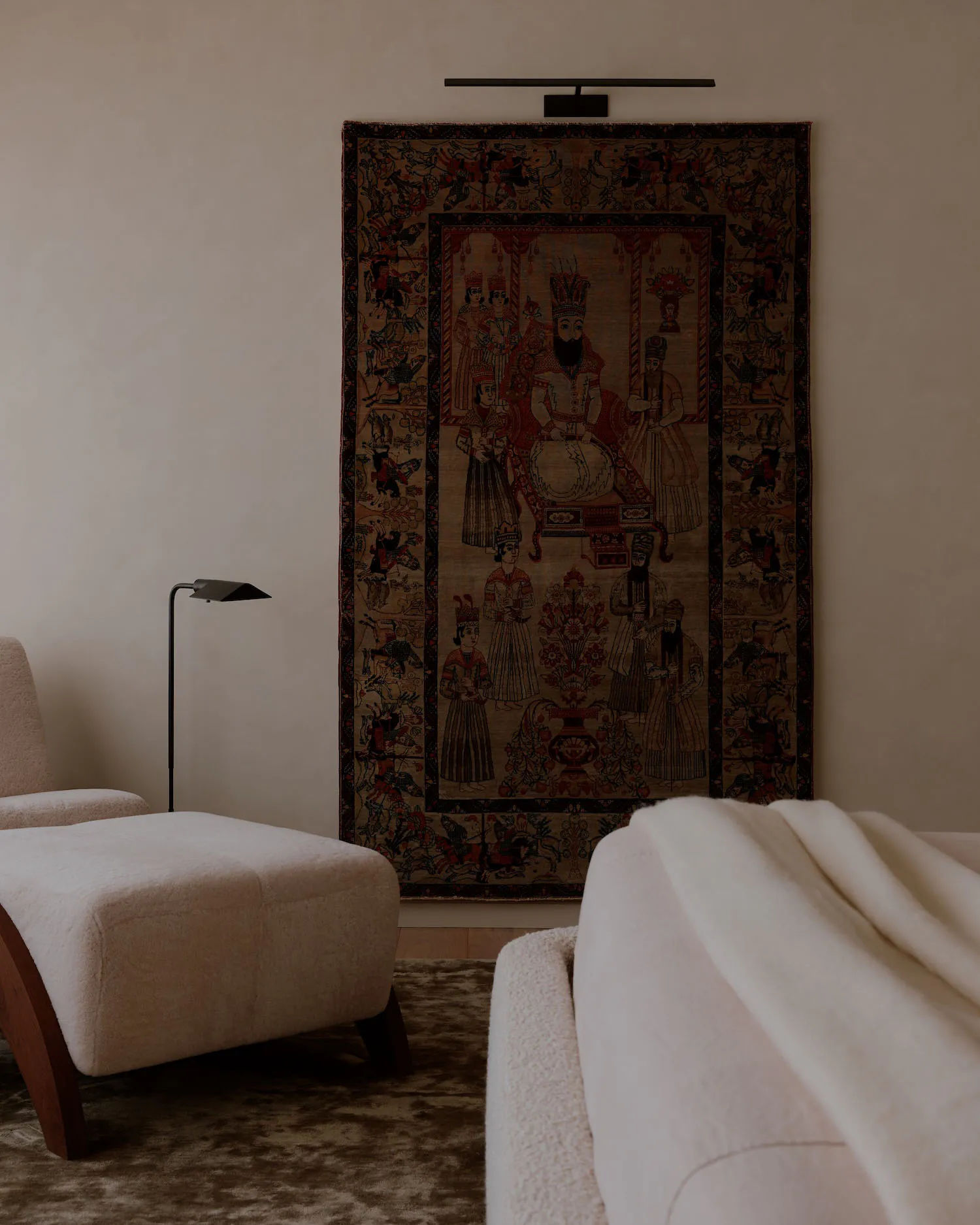 A large antique Persian tapestry depicting a royal court scene hangs on a beige wall above a modern black picture light, next to a cream bouclé armchair and ottoman in a minimalist living room.