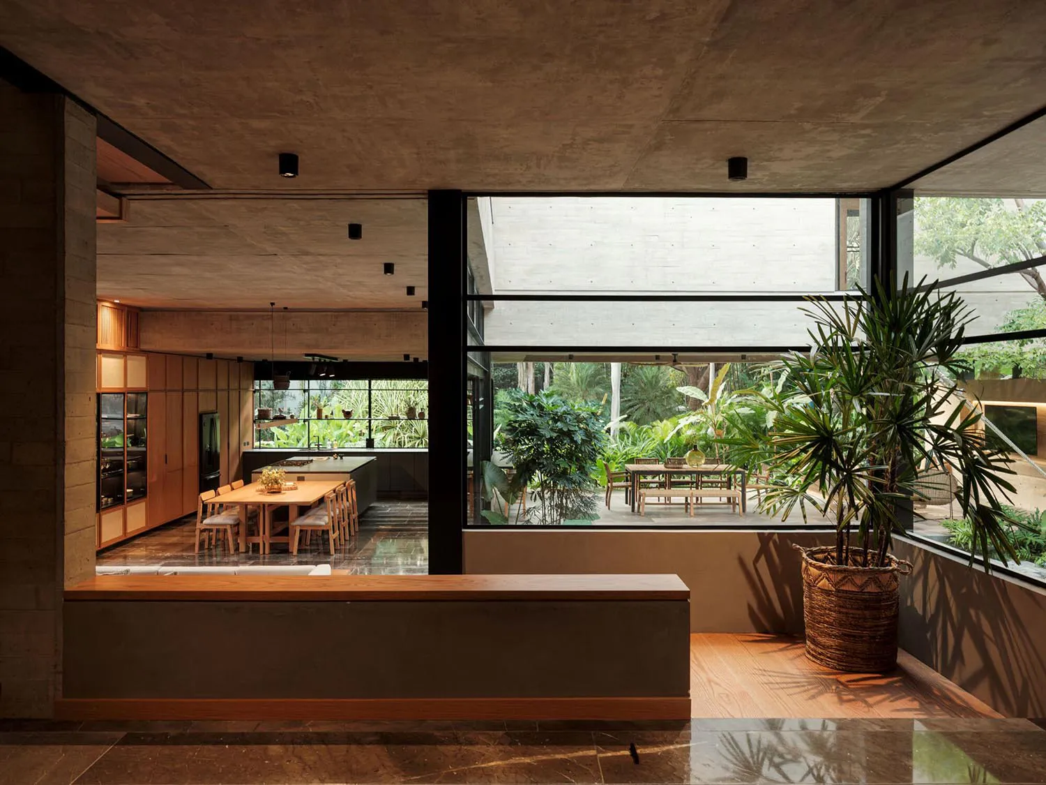 Modern open-plan kitchen and dining area with concrete ceiling, wooden cabinetry, large glass walls overlooking a lush tropical garden and patio.