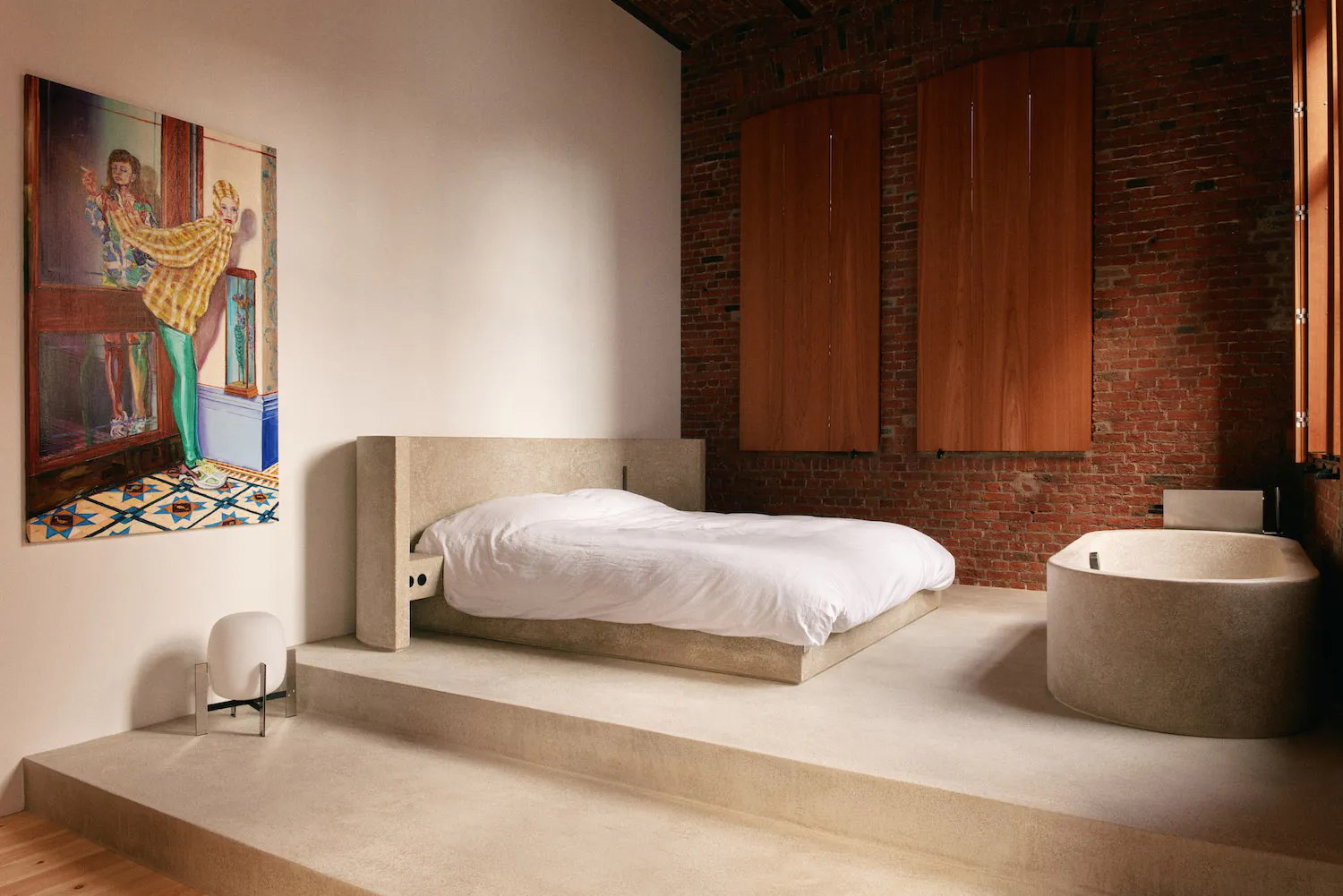 Modern industrial-style bedroom featuring a raised concrete platform bed with white linens, a large freestanding concrete bathtub, exposed red brick walls, wooden wardrobe cabinets, and a colorful contemporary painting on the white wall.