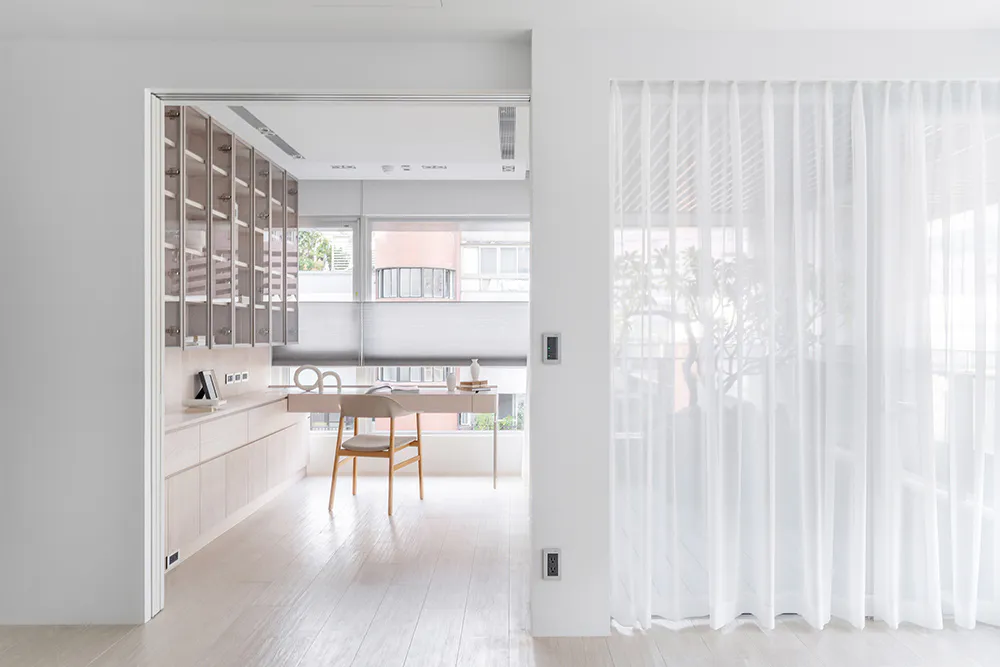 Modern minimalist home office featuring light wood built-in storage, a wooden desk and chair, large windows with roller blinds, and sheer white curtains on the right.