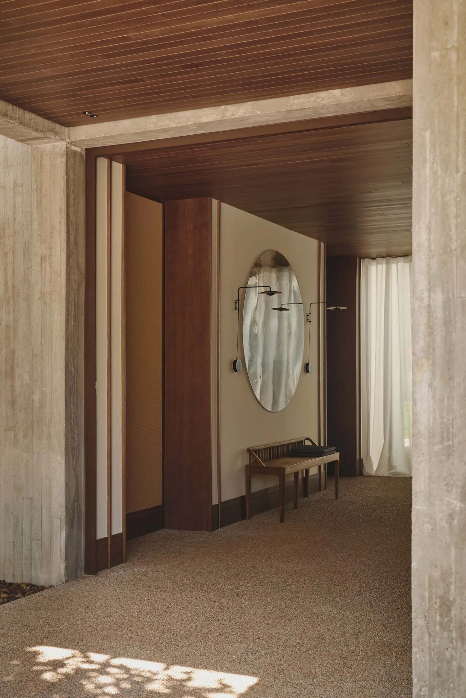 Modern minimalist entryway featuring raw concrete walls, warm wood paneling and ceiling, a large oval mirror with wall sconces, and a wooden bench on a textured carpet floor.