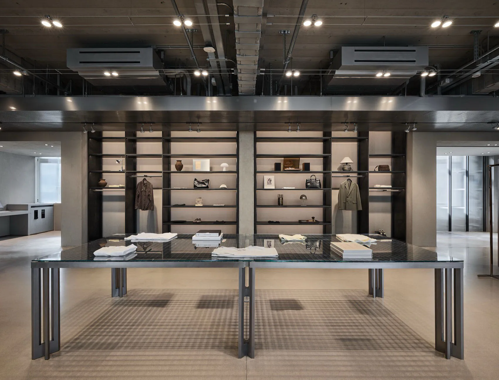 Modern industrial-style luxury clothing boutique interior featuring exposed concrete ceiling, dark steel shelving units, and a large glass-top display table in the foreground.