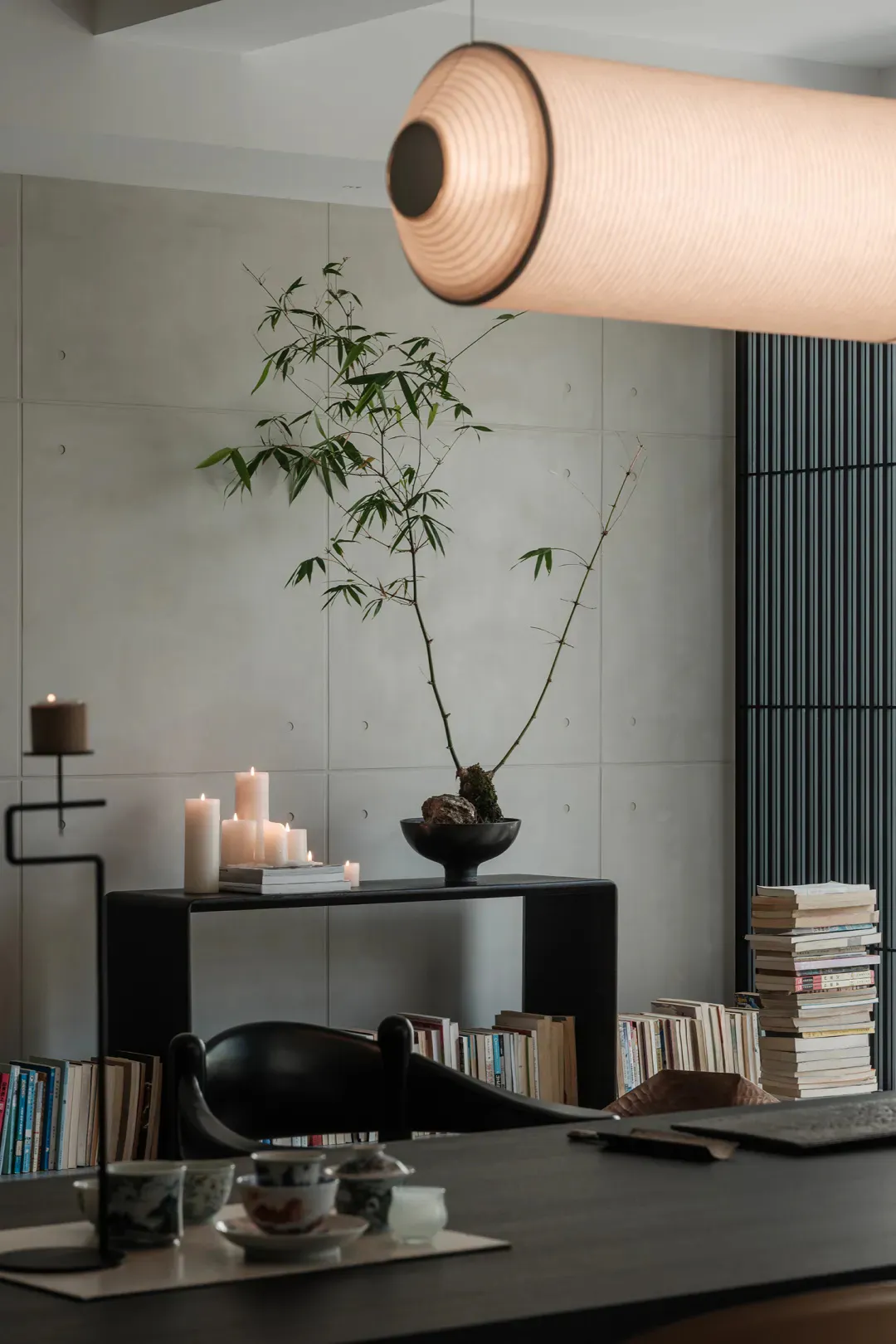 Modern minimalist dining area with dark wood table, black chair, concrete wall, bamboo plant in black bowl, lit candles, stacked books, and a large cylindrical pendant lamp.