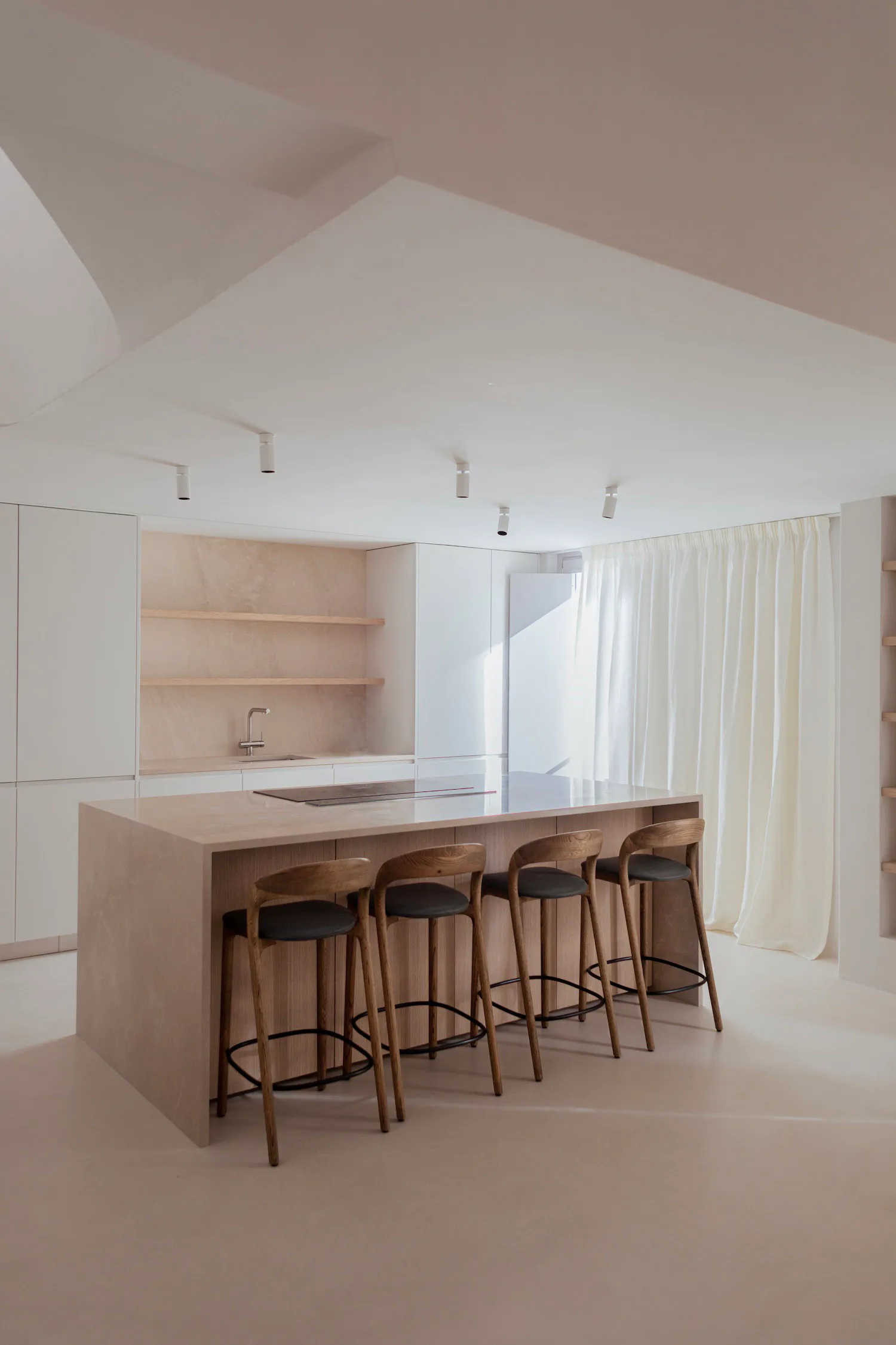 Modern minimalist kitchen with a large beige stone island, integrated cooktop, wooden bar stools with black seats, light oak cabinetry, and sheer white curtains in a bright, airy space.