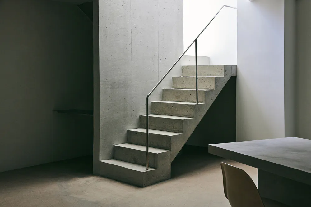 Minimalist interior featuring a raw concrete staircase with thin metal handrail and matching concrete dining table.