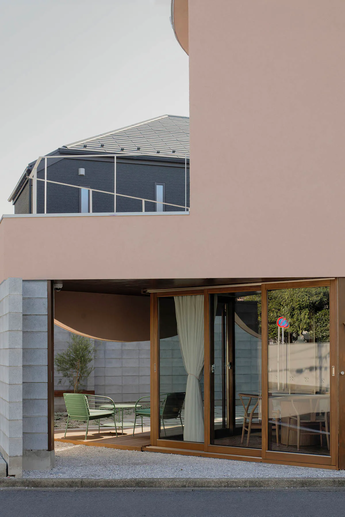 Modern residential building with a soft pink facade, large wooden-framed glass sliding doors, and an outdoor patio furnished with green metal chairs and a small table, next to a dark brick building in the background.