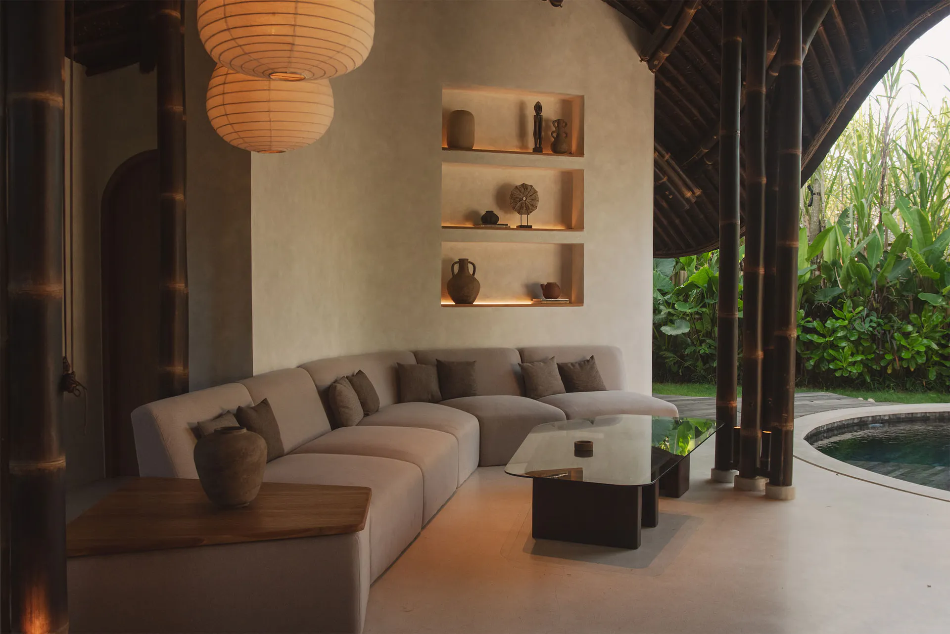 A contemporary open-air living room in a Bali villa featuring a large L-shaped sectional sofa, glass coffee table, built-in illuminated niches with pottery and artifacts, hanging paper lanterns, and dark bamboo structural columns overlooking a curved swimming pool and lush tropical garden.