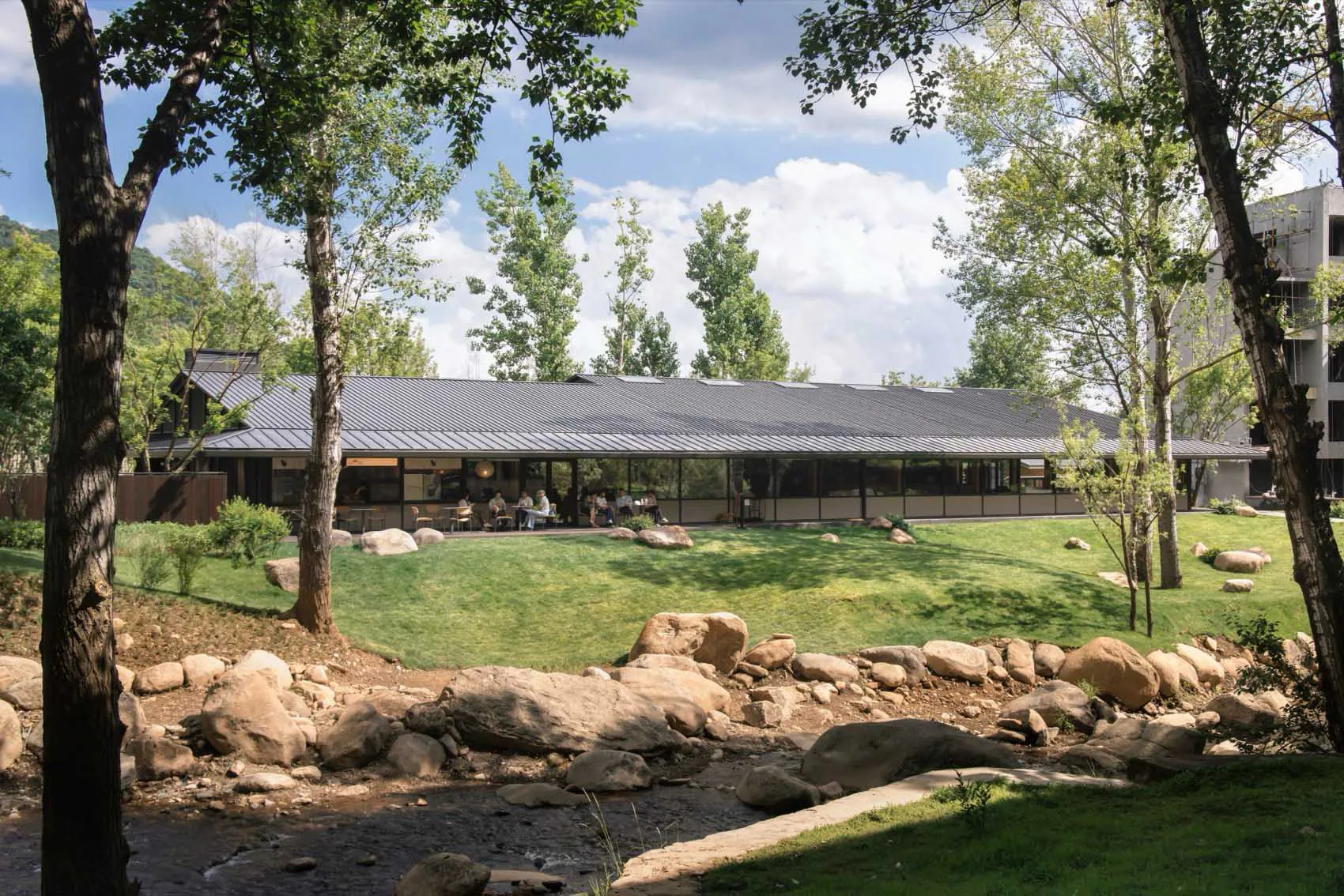 Modern single-story restaurant building with dark corten steel roof and full-height glass walls, situated beside a rocky stream and surrounded by lush trees and lawn.