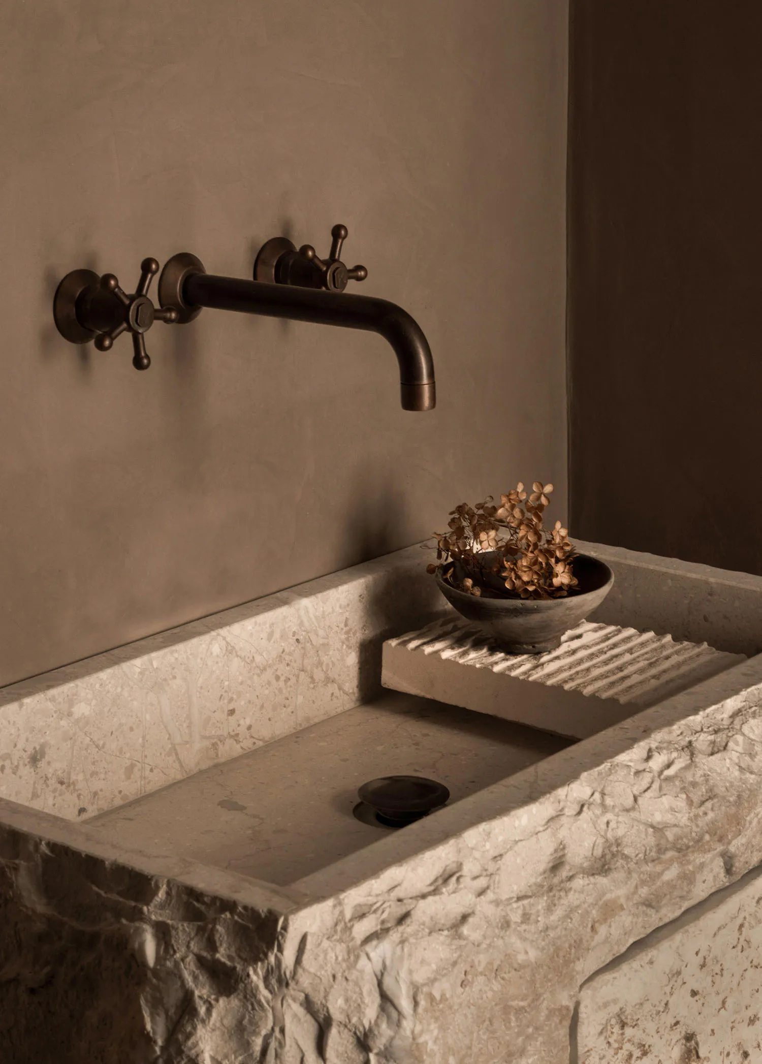 Close-up of a rustic travertine utility sink with a dark bronze wall-mounted faucet featuring cross handles, a small ceramic bowl with dried hydrangeas resting on a ribbed stone ledge, set against a textured plaster wall.