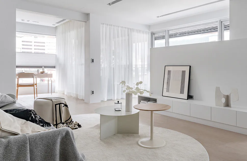 Modern minimalist living room with white curved nesting coffee tables, round white rug, low white media console, framed abstract art, sheer white curtains, and a grey textured sofa in the foreground.