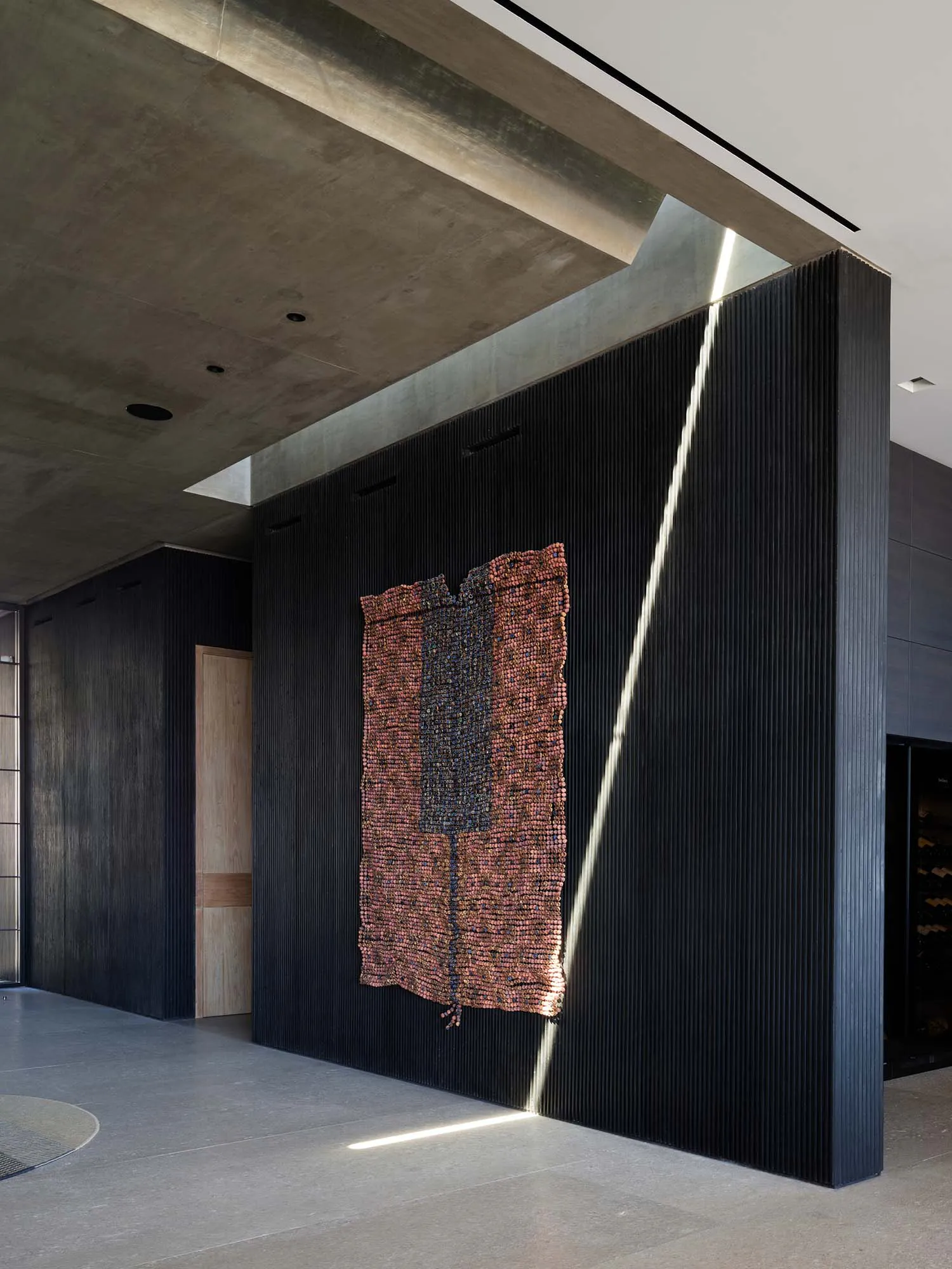 Modern minimalist concrete hallway featuring a large black fluted wall with dramatic linear LED lighting and a vibrant terracotta and black bead tapestry artwork.
