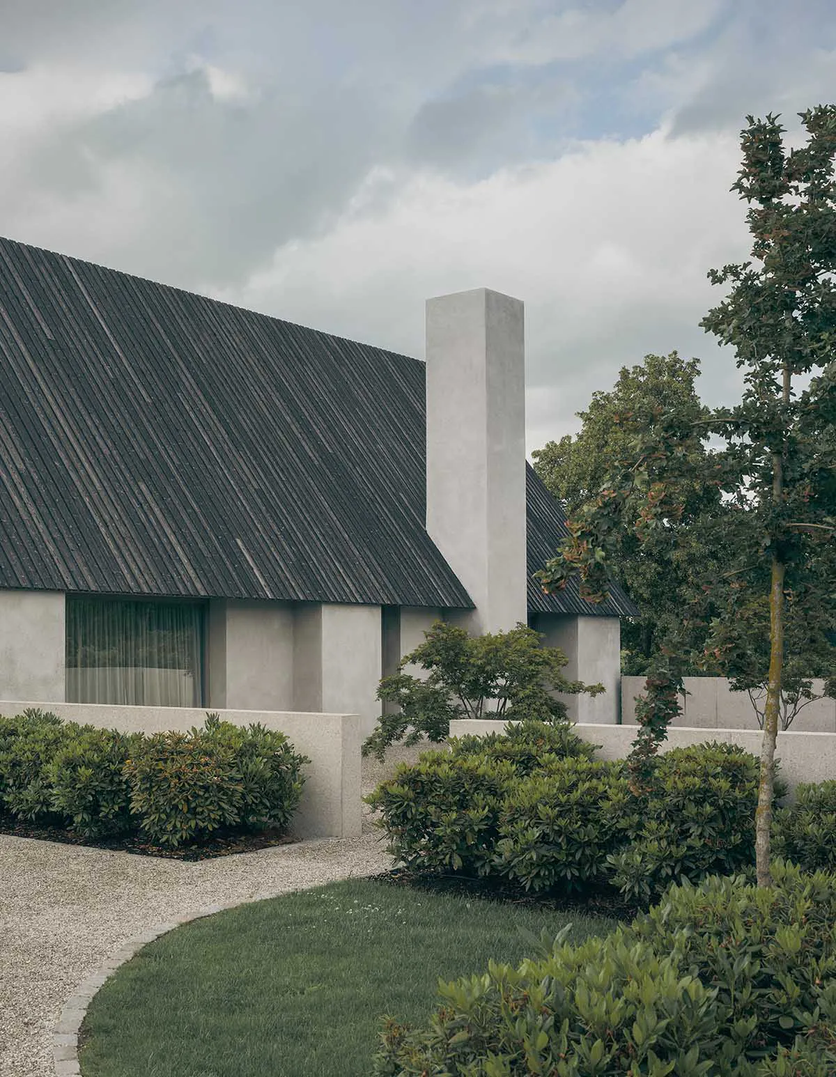 Exterior view of a modern minimalist concrete building with a dark sloped roof, tall chimney, and landscaped courtyard garden featuring manicured hedges, gravel paths, and mature trees under a cloudy sky.