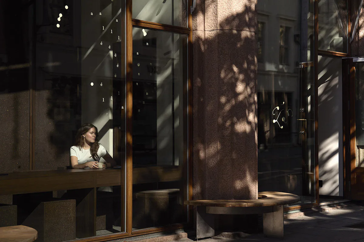 A woman with long brown hair wearing a white t-shirt sits at a wooden counter inside a modern cafe, holding a white coffee cup and looking out the window. Outside, a curved wooden bench sits beside a large textured stone pillar under strong sunlight that casts intricate tree shadows across the glass facade.