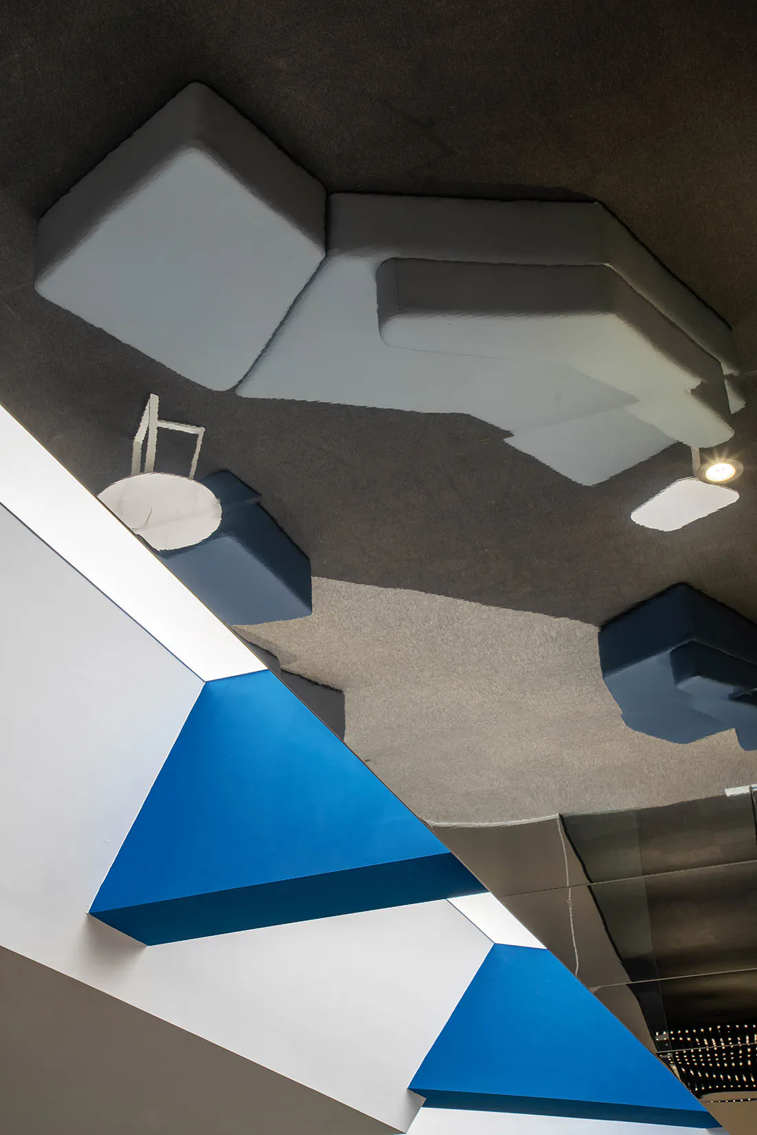 View looking up at a modern geometric ceiling installation made of white, light grey, dark blue, and bright blue felt acoustic panels, with integrated lighting and a small white table and chair visible on the upper level.