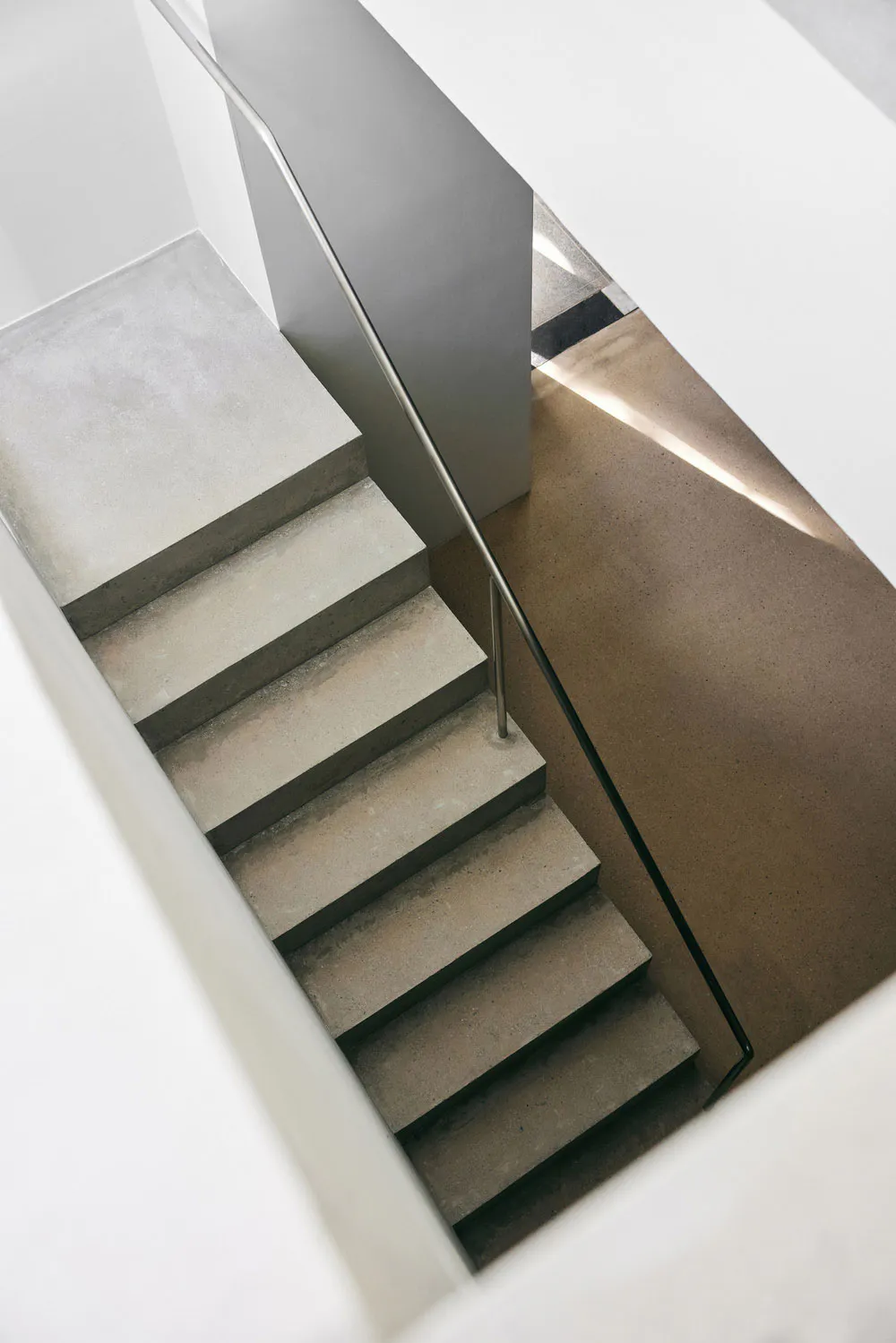 High-angle view of a minimalist concrete staircase with stainless steel handrail and dramatic natural light casting long shadows on the lower landing.