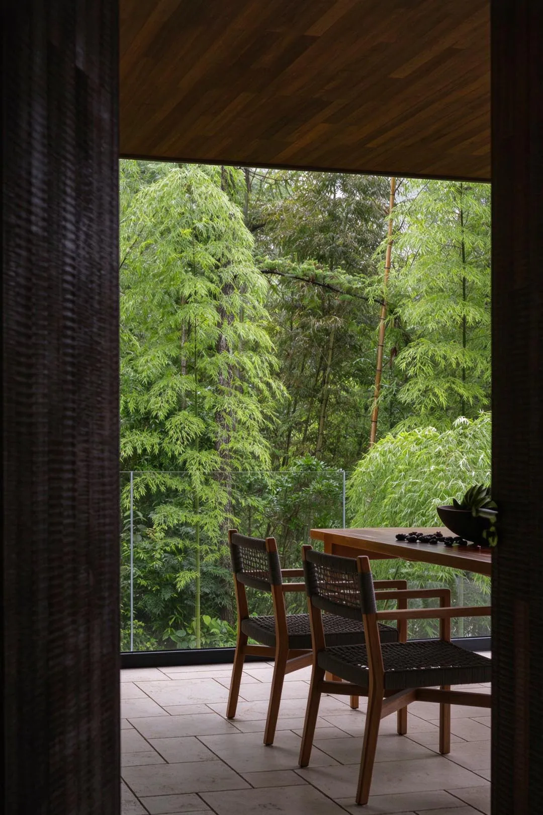View from inside a wooden structure looking out onto a tiled balcony with teak dining chairs and table, overlooking a dense bamboo and conifer forest.