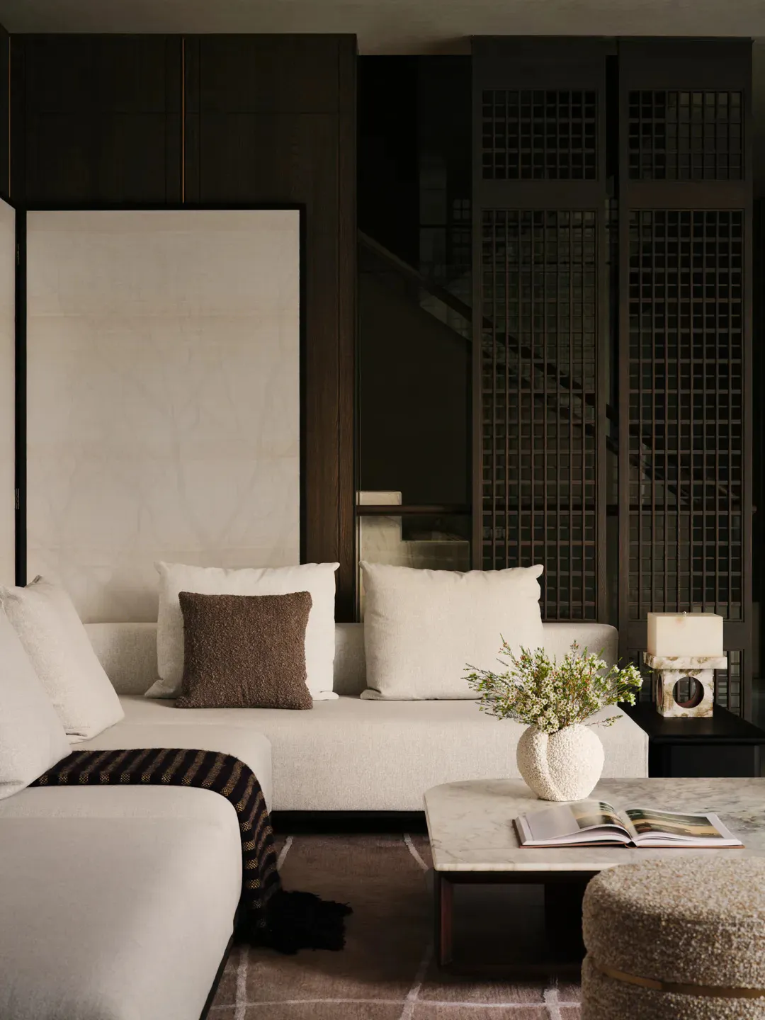 Modern minimalist living room with a light-colored L-shaped sectional sofa, brown textured throw pillow, dark wood cabinetry, marble coffee table, and a staircase visible through a lattice screen in the background.