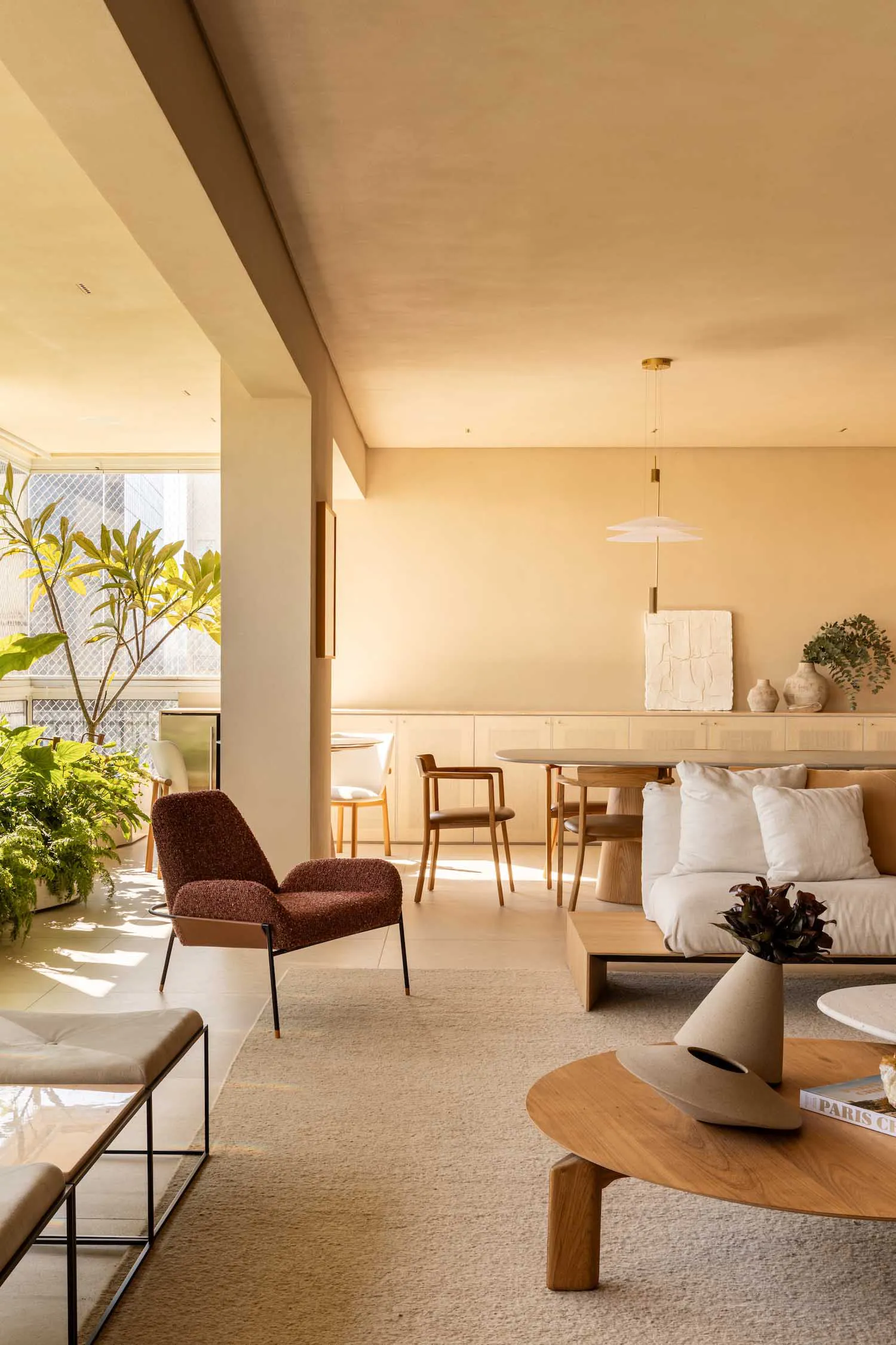Modern minimalist living room featuring a curved oak coffee table, rust boucle armchair, neutral sofa, pendant light, and abundant indoor plants under warm sunlight.