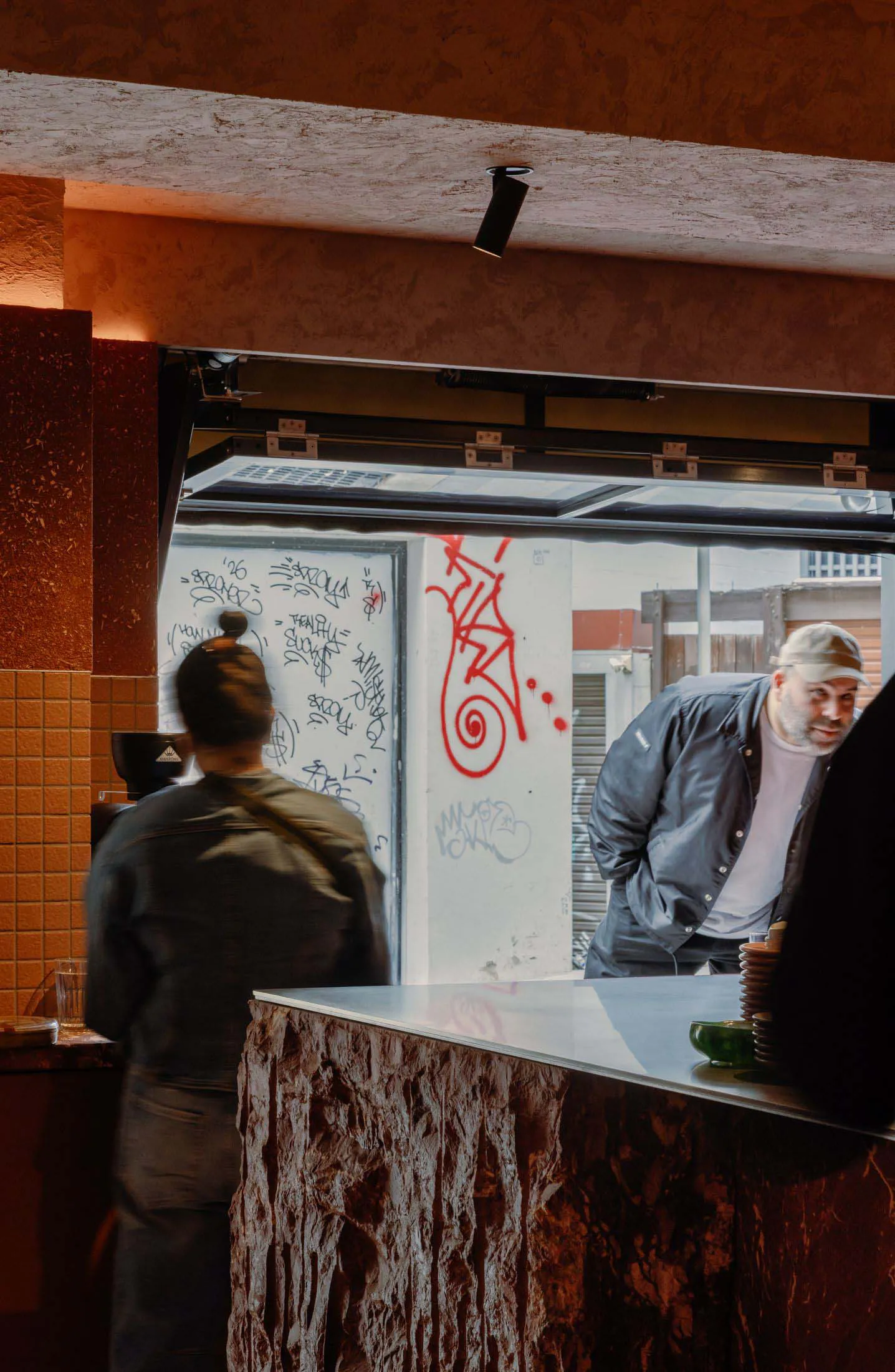 Interior view of a modern cafe showing a textured stone service counter, a barista with hair in a bun facing away, and a bearded man in a cap leaning on the counter. The background features white walls covered in colorful graffiti and red street art, with warm orange lighting and an open pass-through window.