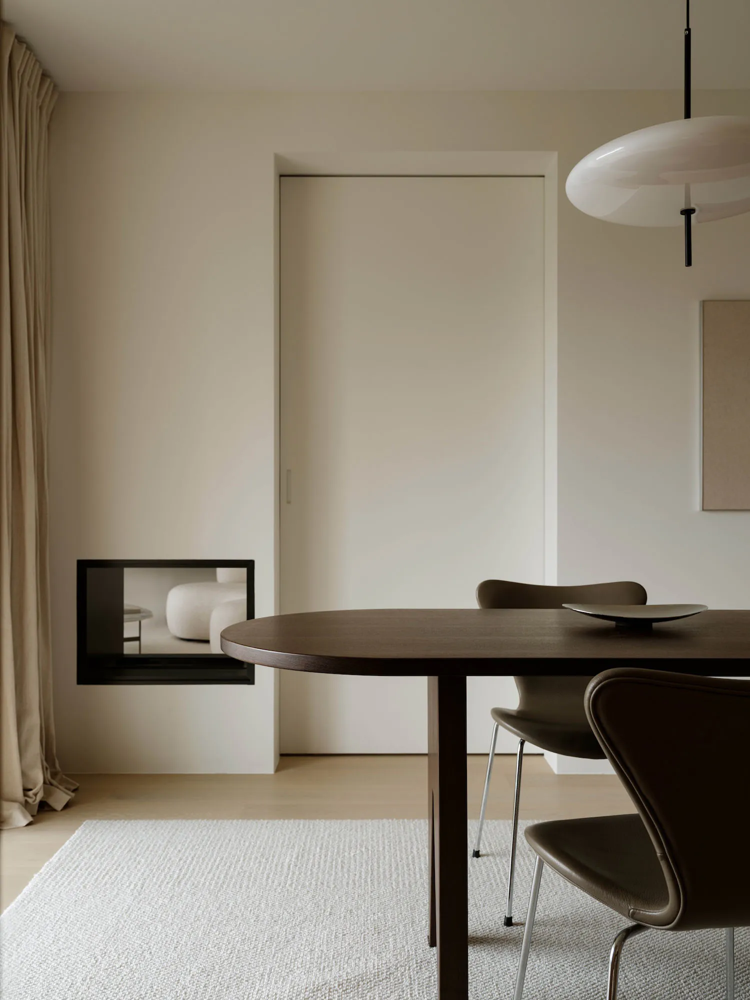 Modern minimalist dining area featuring a dark walnut round table, brown leather chairs, white textured rug, and a large white pendant lamp in a neutral beige interior.