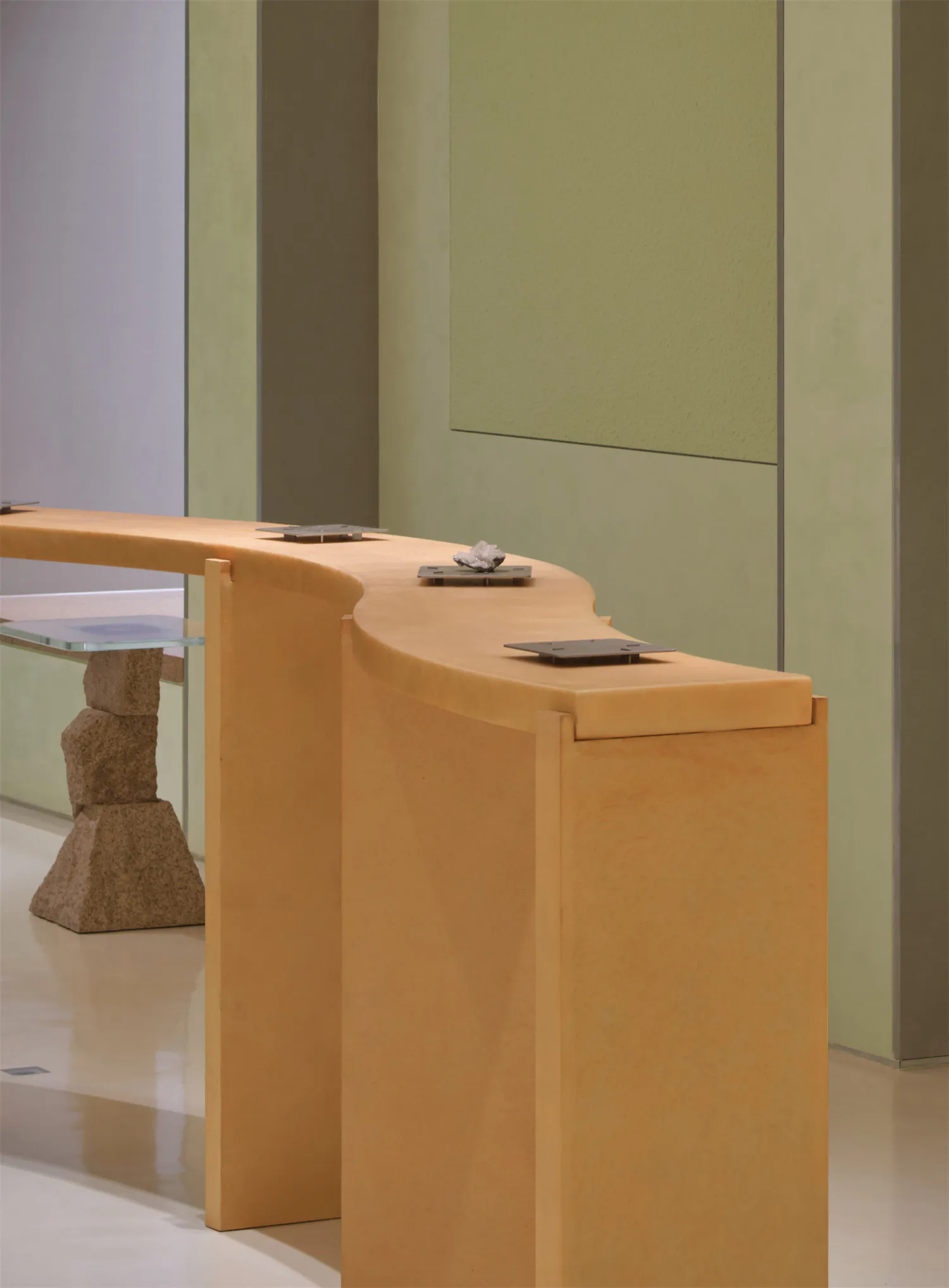 Modern curved light-wood reception desk with stacked stone pedestal and glass side table against sage green and beige walls.