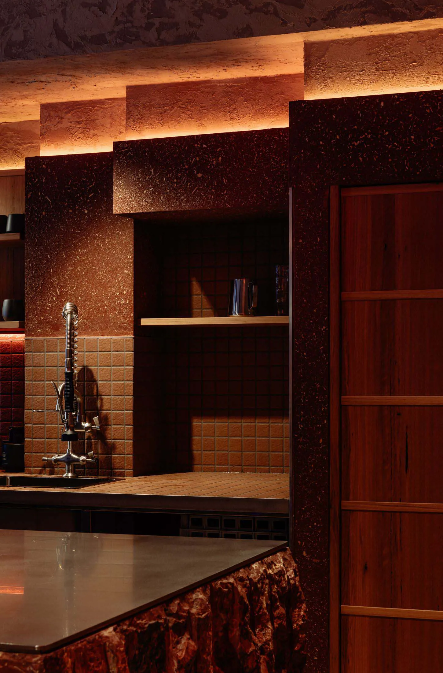 Modern kitchen featuring dark red terrazzo walls and bar counter, wooden cabinetry, brown tile backsplash, industrial-style faucet, and warm ambient lighting from above.