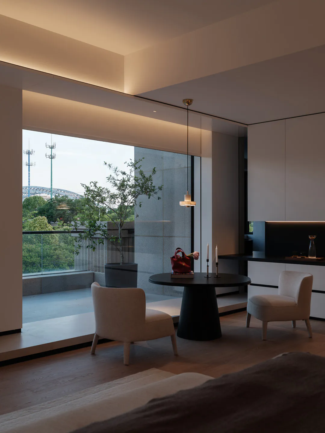Modern minimalist living and dining area with two beige upholstered chairs, a round black pedestal table, large floor-to-ceiling windows overlooking green hills and city infrastructure, light oak flooring, and warm ambient ceiling lighting.