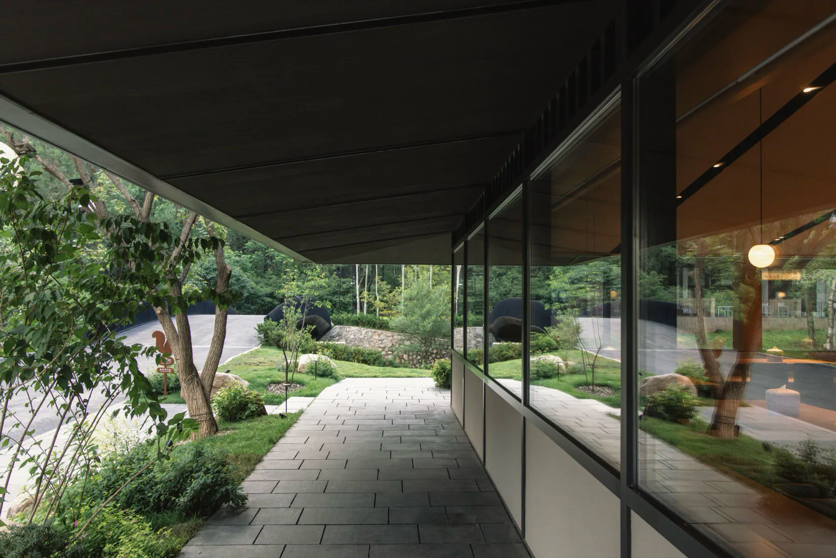 Modern covered veranda with dark wood ceiling, large glass windows, and a stone-paved walkway leading into a lush green courtyard garden with trees and rocks.