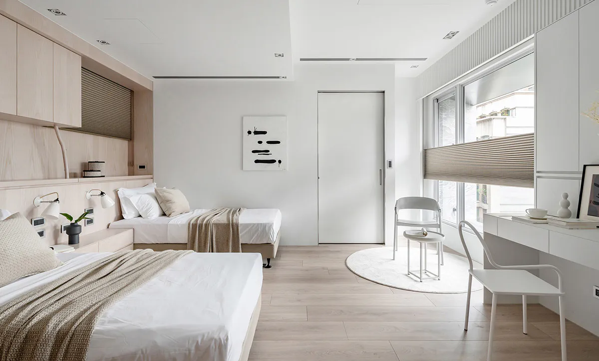Modern minimalist bedroom interior with two single beds, light wood cabinetry, white walls, a built-in desk with white chairs, and large windows with neutral blinds.