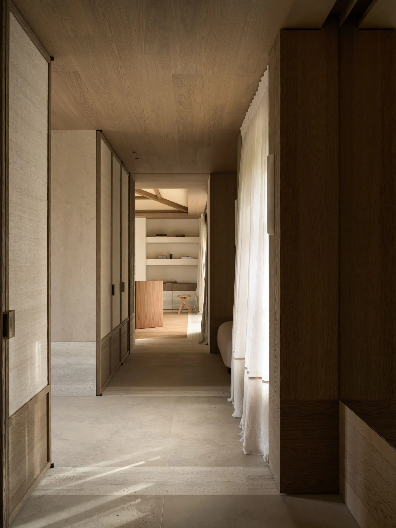 A long, minimalist hallway featuring light wood ceilings and wall paneling, polished concrete floors, sheer white linen curtains, and a view into a study area with built-in shelves.