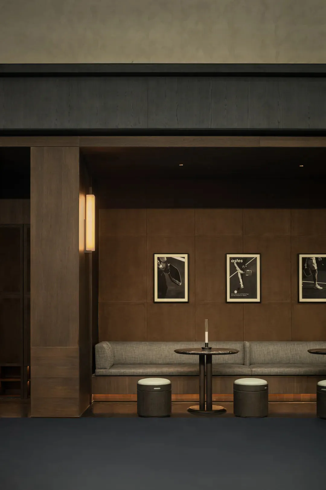 Modern lounge interior with dark wood paneling, a gray upholstered banquette, round marble-top table, cylindrical stools, vertical wall sconce, and three framed black-and-white pickleball photographs on the wall.