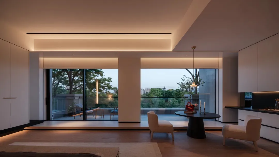 Modern minimalist open plan living and dining room with white upholstered chairs, black round dining table, warm cove lighting, and large glass windows overlooking trees and city skyline at dusk.