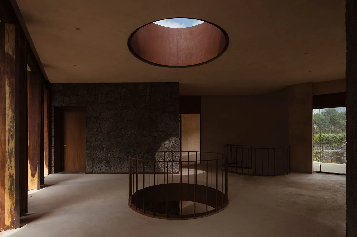 Empty modern brutalist interior featuring a large circular skylight, exposed concrete, dark volcanic stone wall, and a cylindrical metal railing in the center.