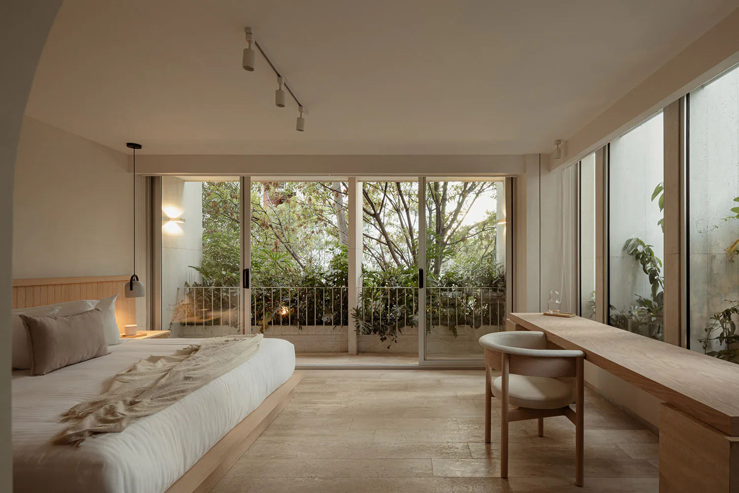 Modern minimalist bedroom featuring a low platform bed with beige linens, a long oak wood desk with a curved upholstered chair, large sliding glass doors revealing a lush green garden, and warm track lighting.