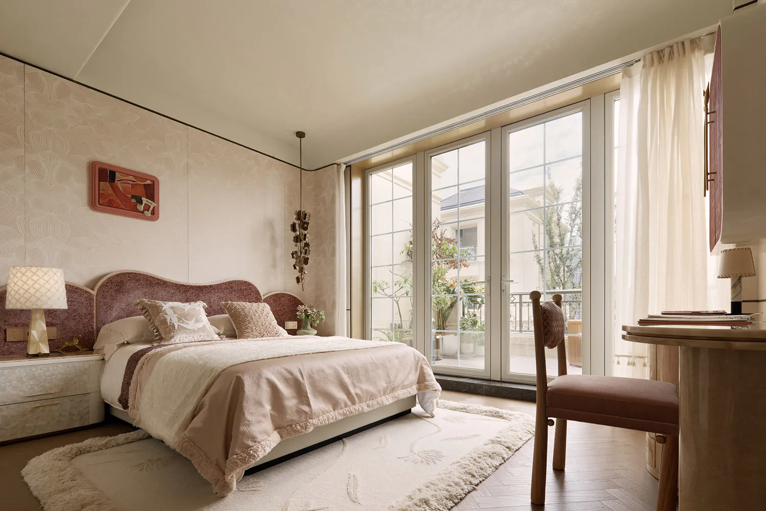 Elegant bedroom featuring a curved dusty-rose velvet headboard, neutral textured walls, cream bedding, wooden desk and chair, and large glass doors leading to a balcony with potted plants.
