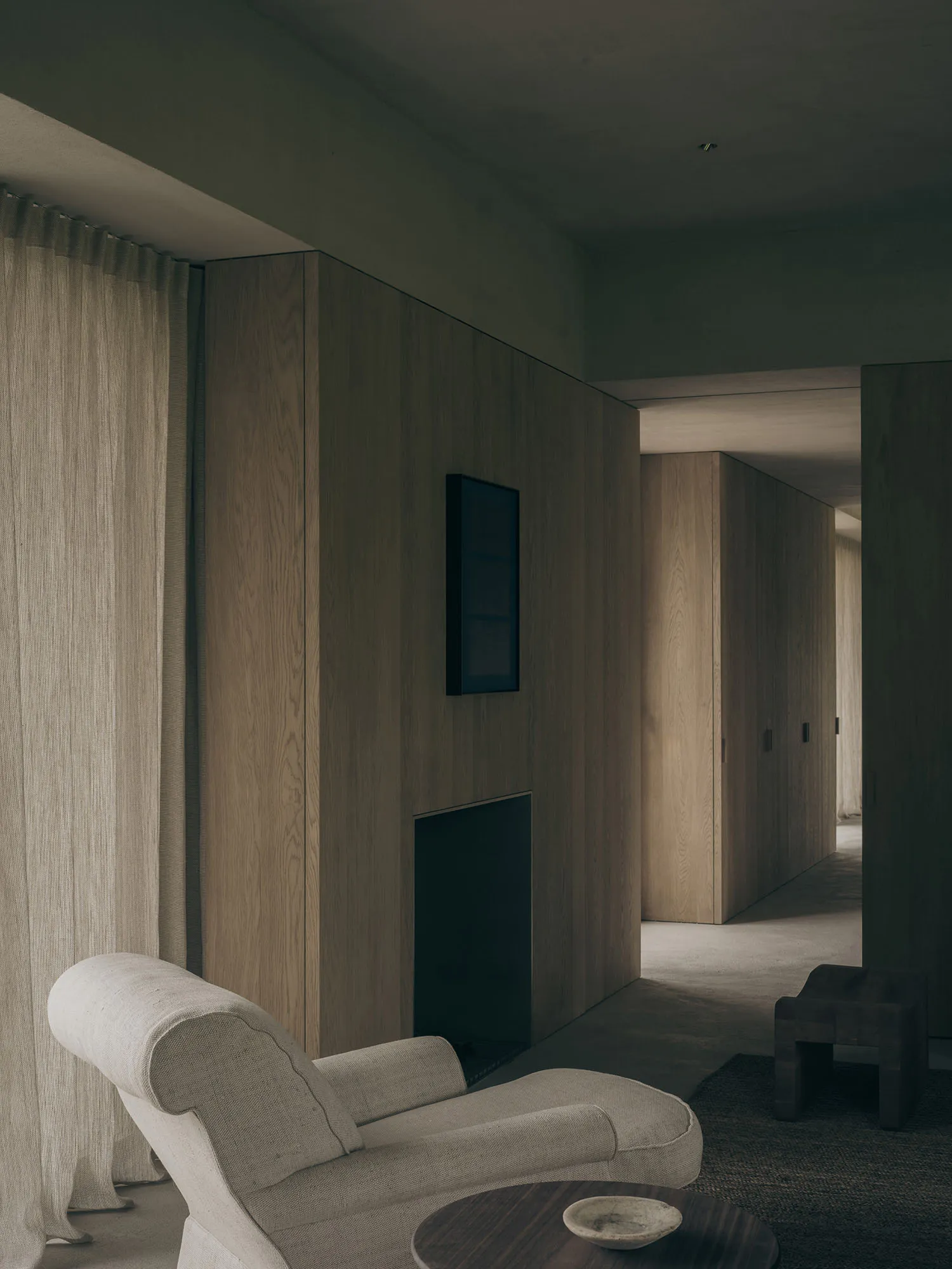 A contemporary minimalist living room featuring light oak wood paneling, a sculptural off-white armchair, a dark wooden side table, and a recessed fireplace niche under soft ambient lighting.