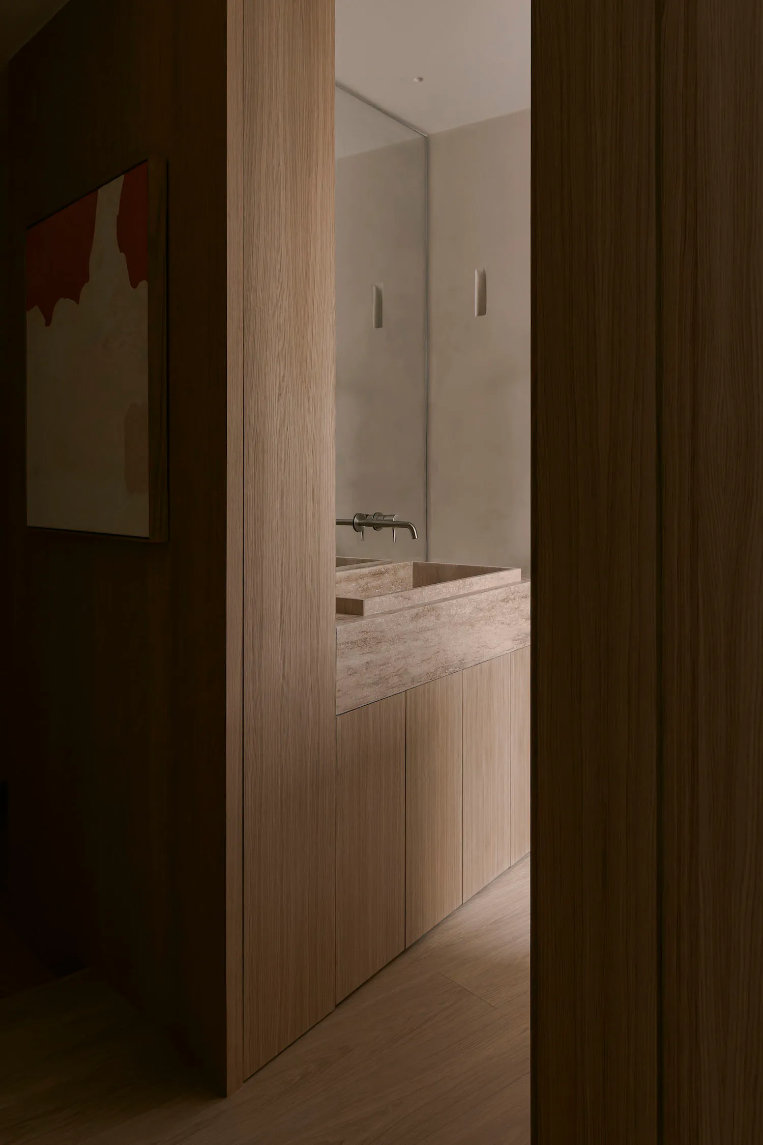 View through a wooden doorway into a modern minimalist bathroom with a long travertine stone vanity, wall-mounted faucet, large mirror, and warm wood cabinetry.