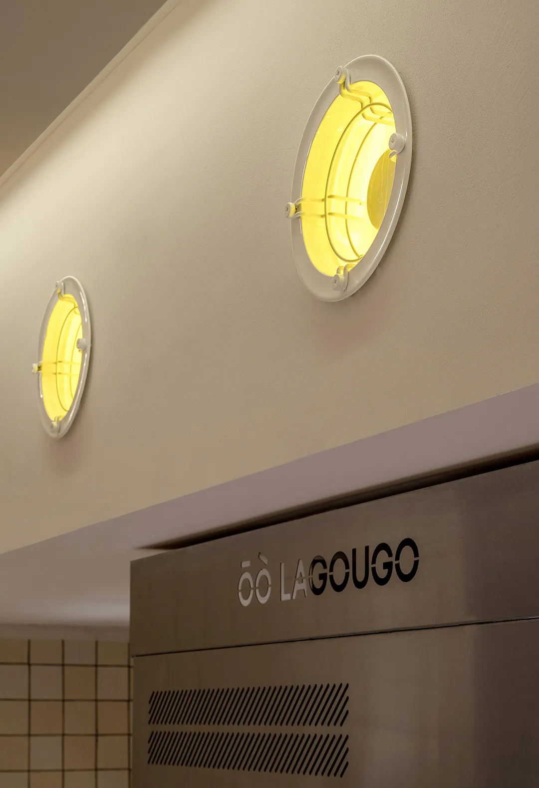 Two circular yellow-glowing porthole wall lights mounted on a beige wall above a stainless steel range hood with the brand name LAGOUGO.