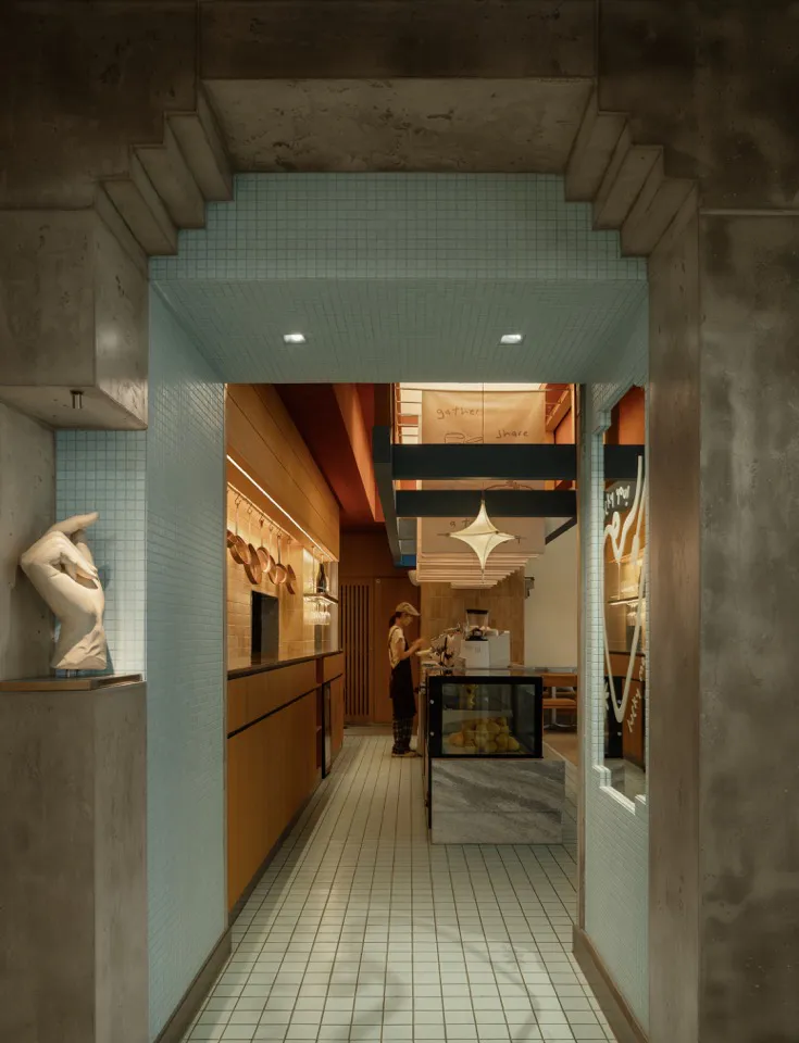 View through a concrete archway into a modern cafe featuring light blue tiled walls, warm wood cabinetry, a marble and terrazzo display counter, and a staff member preparing items in the background.