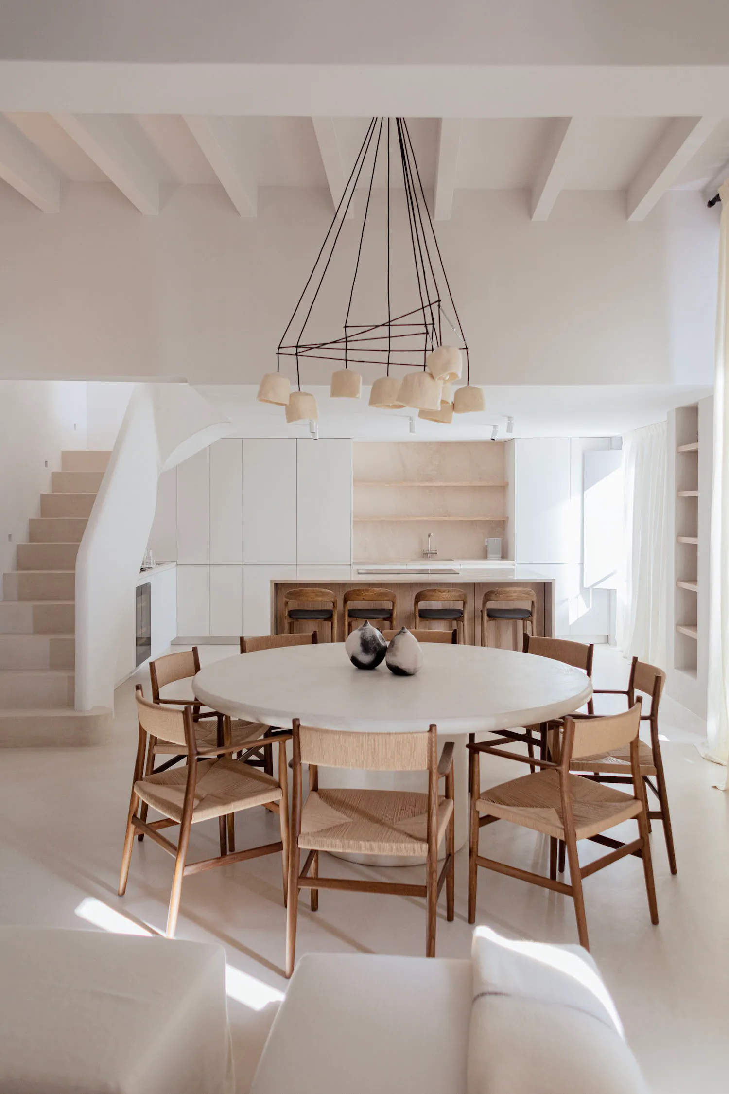 Bright minimalist dining area featuring a large round white marble table, six oak chairs with woven seats, a dramatic black geometric chandelier, white walls, and a view of the kitchen and staircase in the background.