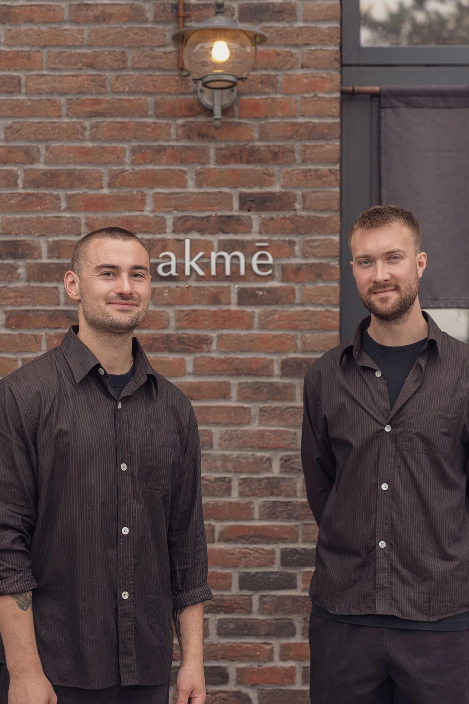 Two men with short hair and light stubble wearing dark pinstriped button-down shirts standing in front of a red brick wall with the sign 'akmē' and a lit wall lantern.