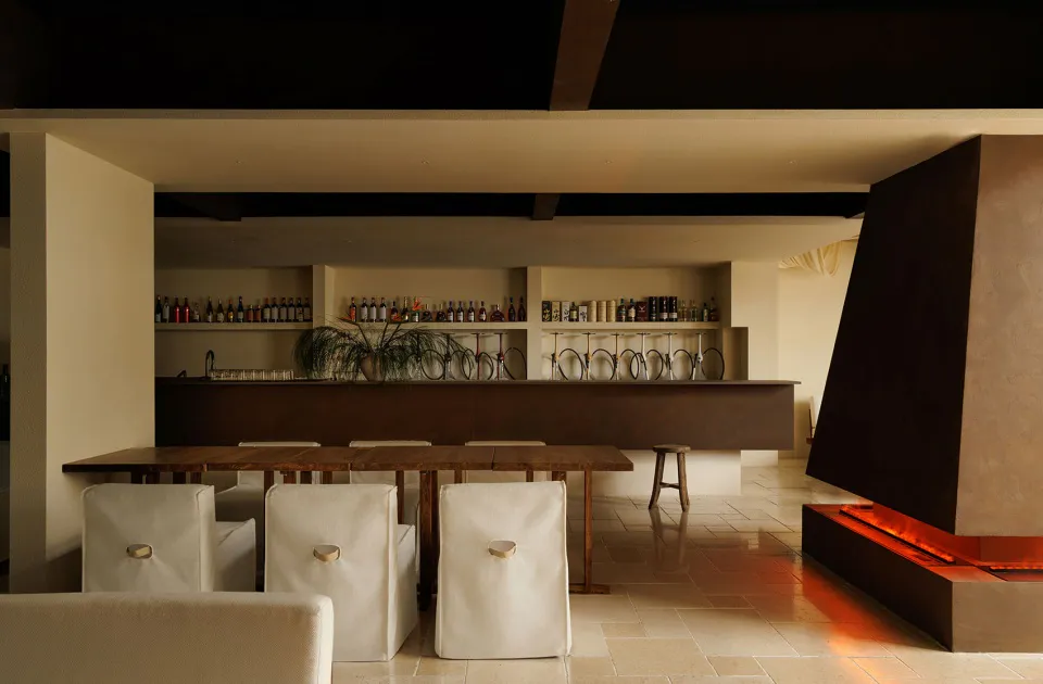 Modern minimalist interior featuring a long wooden dining table with white upholstered chairs, a dark wood bar counter with wine glass racks and liquor shelves, and a large concrete fireplace with glowing fire on the right.